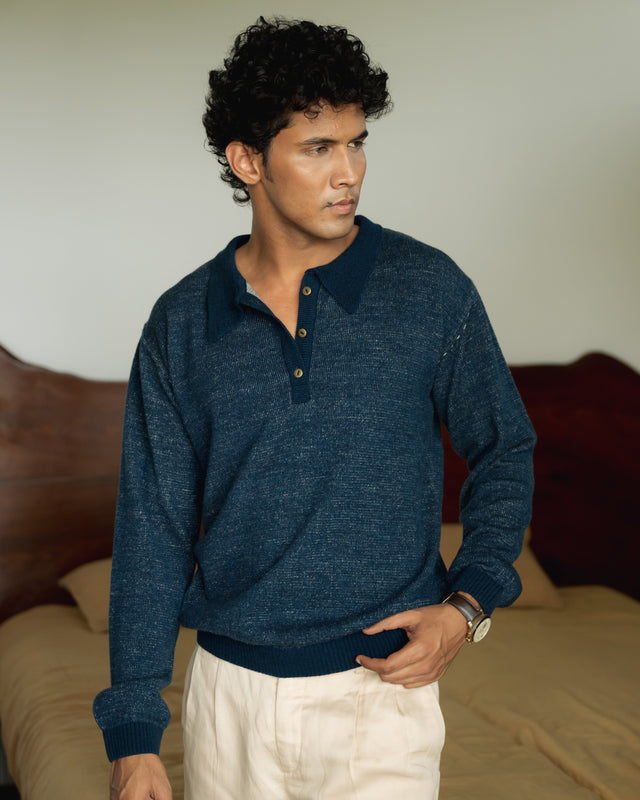 Man wearing a blue sweater and white pants standing in a bedroom.