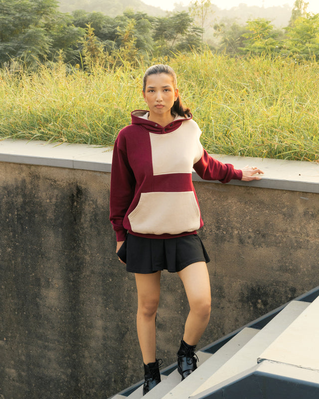 Woman in a red and beige hoodie and black skirt standing on a concrete ledge with greenery in the background