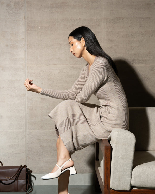 Woman sitting on a couch wearing a gray dress and white shoes, with a brown handbag beside her.